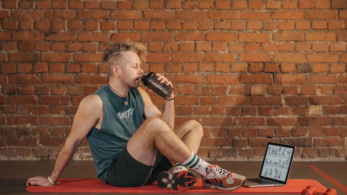 Athletic man sitting on exercise mat with water bottle, taking a break from workout, in a rustic indoor setting.